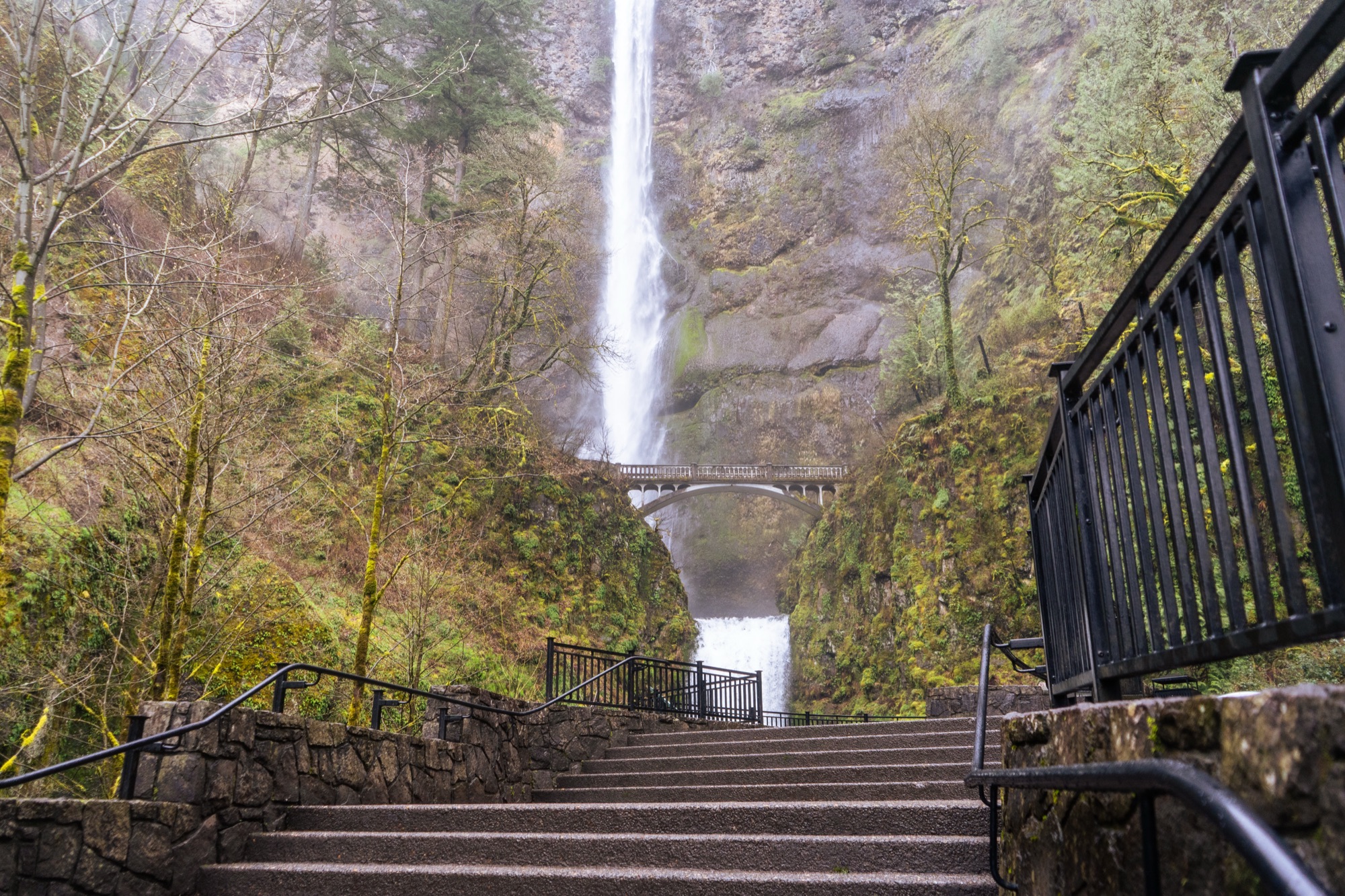Stone steps and a footbridge beneath a tall waterfall.